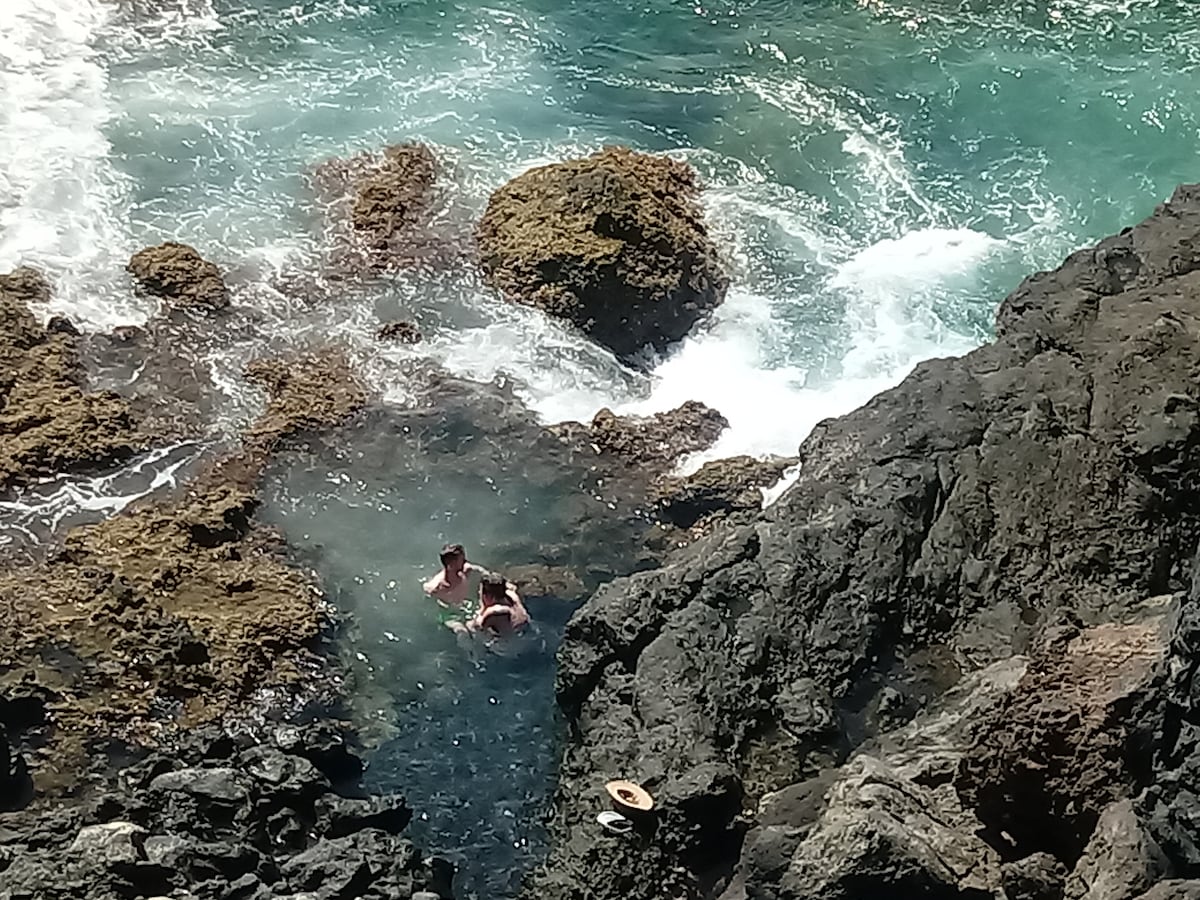 Clear turquoise water surrounds rocky formations, creating a natural pool. Two individuals are visible enjoying the shallow water, which is gently lapping against the rocks. The contrast of the dark stones and the bright sea is prominent, highlighting the beauty of the coastal landscape.