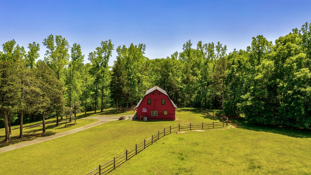 A red barn is positioned within the expansive landscape, surrounded by lush greenery. Trees border the property, and a gentle path leads to the barn, which has a distinctive triangular roof and several large windows reflecting natural light.