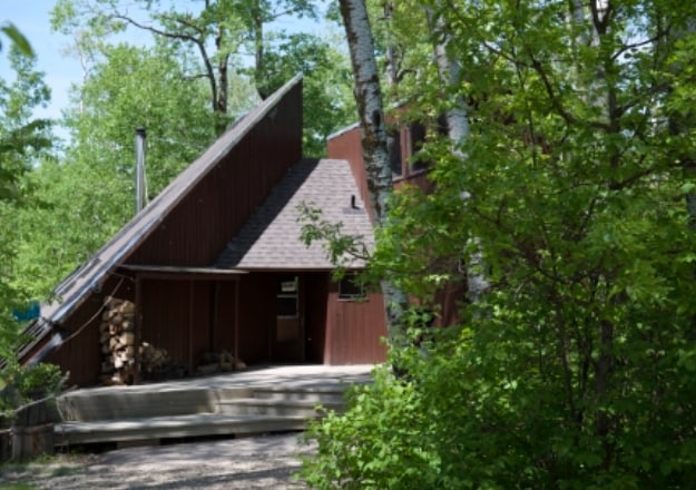 A rustic cabin is nestled among trees, featuring an angled roof and wooden exterior. A deck extends from the cabin, offering a space for relaxation. Stacked firewood is visible beside the entrance, surrounded by lush greenery that enhances the secluded atmosphere.