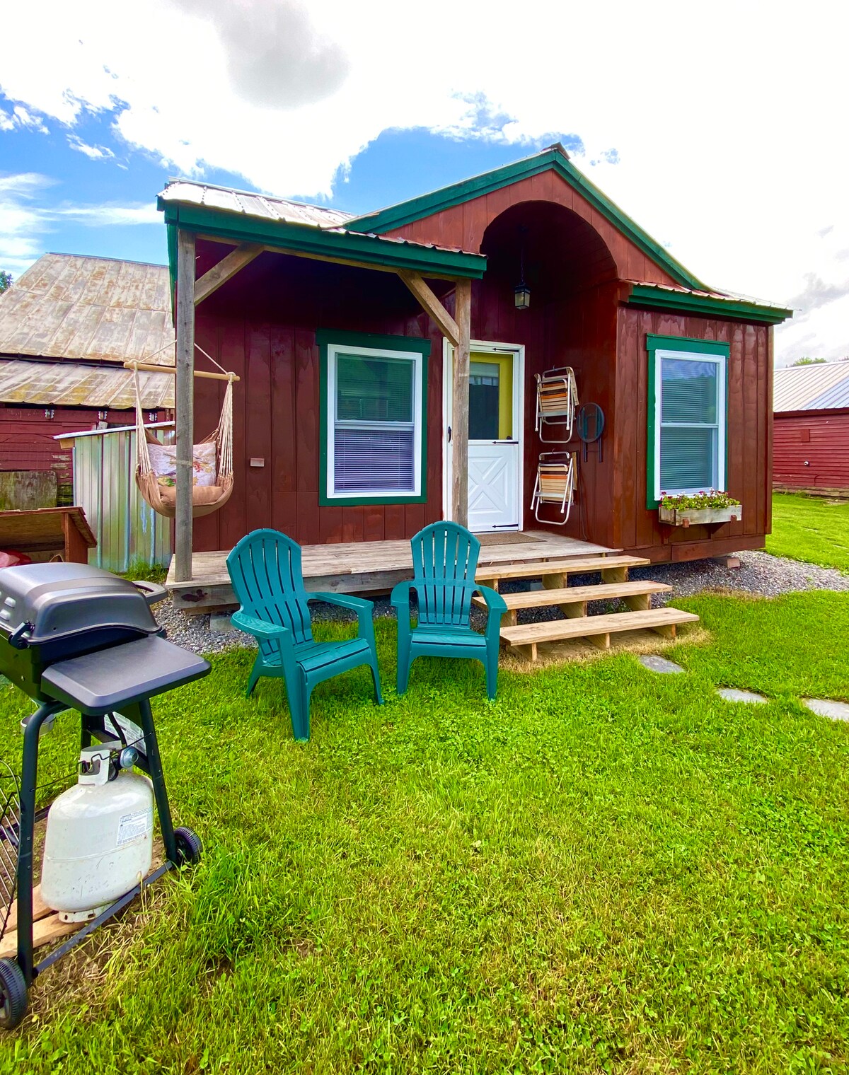 The exterior of a tiny home showcases a reddish-brown facade with a peaked roof. Two green chairs sit on the front steps, inviting relaxation. A gas grill is positioned nearby on a stone surface, while colorful flower boxes decorate the window, adding a touch of charm.