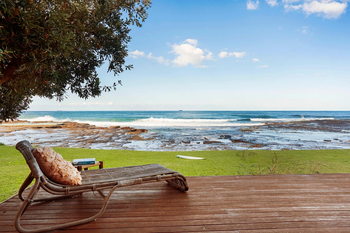A sun-lounging chair sits on a wooden deck, facing the expansive ocean view. The lush green lawn extends towards rocky tidal pools and gentle waves lapping the shore. The sky is clear with a few scattered clouds, creating a serene coastal atmosphere.