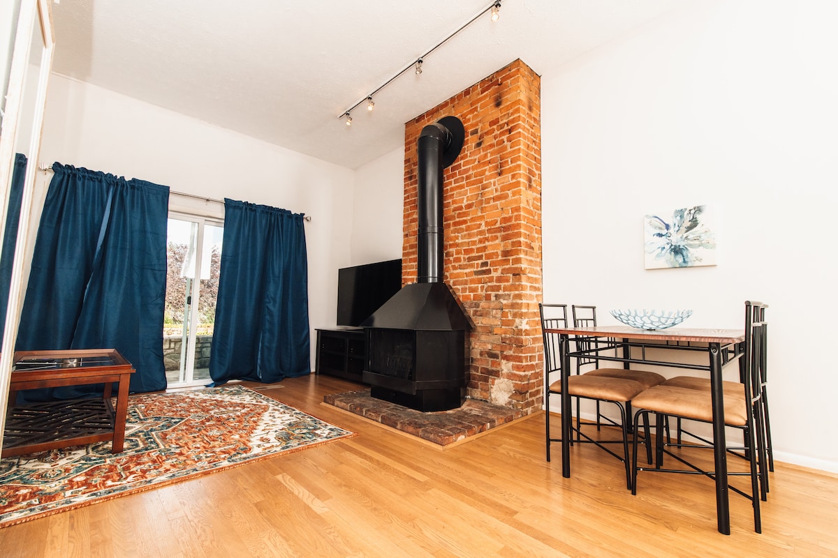 The living area features a cozy wood-burning stove against a brick wall, complemented by a dining table with four chairs. Large blue curtains frame a window that opens to a view outside. A colorful area rug lies beneath a wooden coffee table, adding warmth to the space.