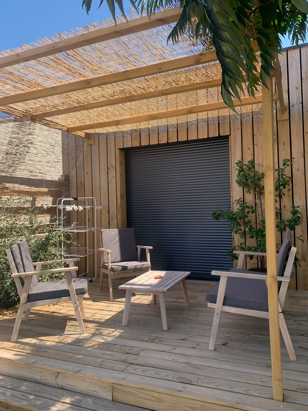 A private wooden terrace features a small table surrounded by four comfortable chairs. The space is partially shaded by a pergola, and greenery is visible along the walls, contributing to a relaxing outdoor atmosphere.