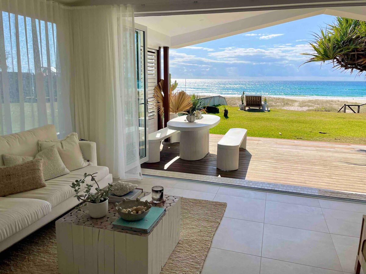 An airy living area features a light-colored sofa and a round dining table set against large glass doors that open to the beach. Natural light floods the space, accentuating the soft, neutral tones of the decor and a view of the ocean beyond.