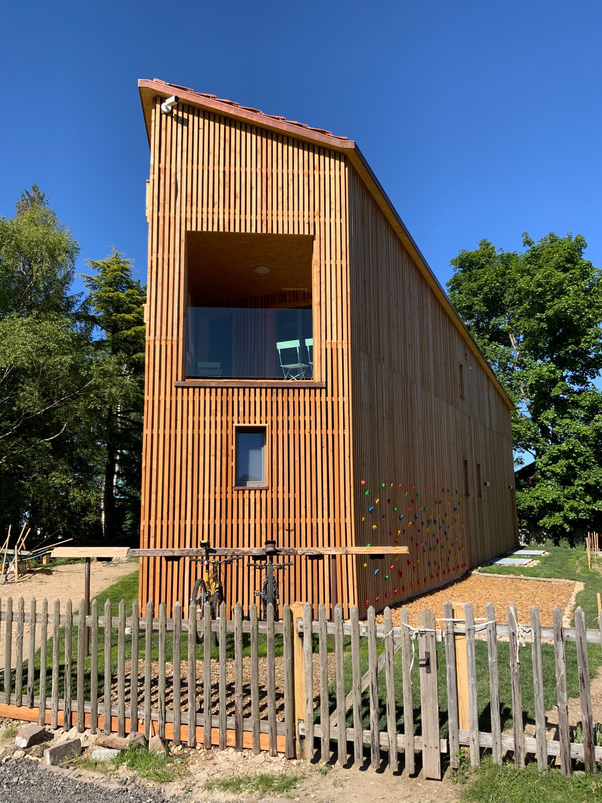 A modern wooden structure is depicted, featuring vertical slats and large windows that provide natural light. A climbing wall is visible on one side, while bicycles are securely mounted. The surrounding greenery and fence enhance the outdoor setting.