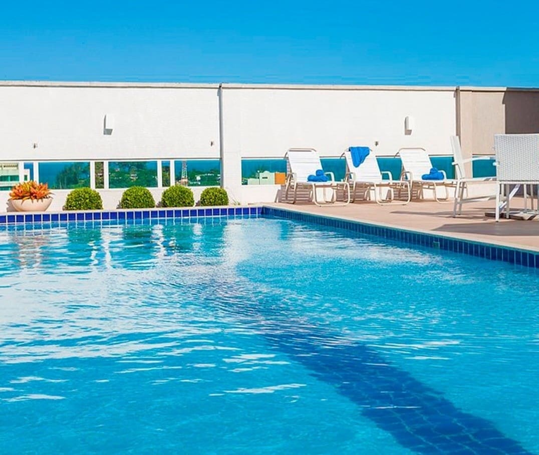 A clear blue swimming pool is visible, reflecting the sky above. Sun loungers are positioned along the poolside, paired with small green shrubs for a touch of greenery. The surrounding area features a light-colored deck and large windows, enhancing the spacious feel.