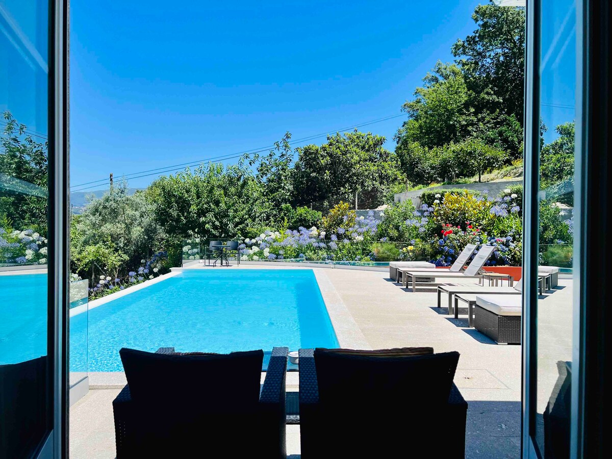 A view of the salt-water pool is visible through expansive glass doors. Surrounding trees and vibrant flowers create a lush garden backdrop. Lounge chairs line the poolside, inviting relaxation in a serene outdoor setting.