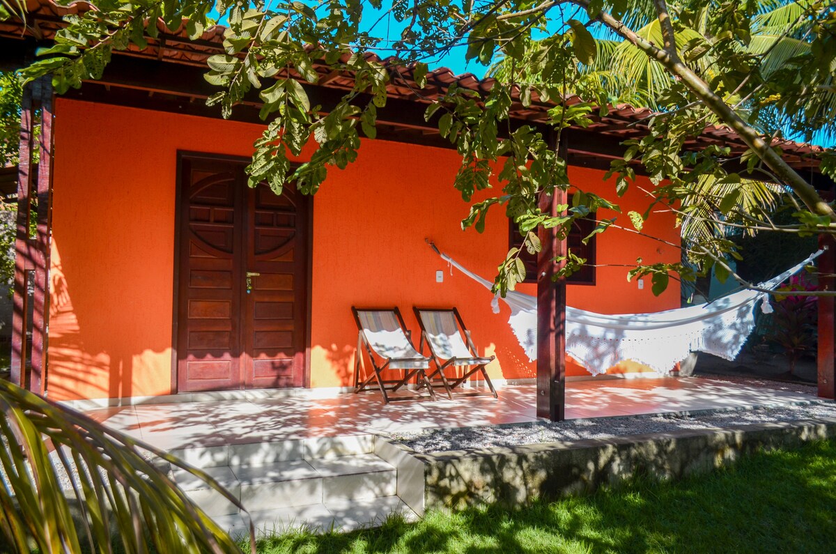 The exterior features an orange façade with a welcoming wooden door. Two lounge chairs are positioned on a covered patio, accompanied by a hammock strung between supports. Lush greenery provides shade and enhances the outdoor living space.