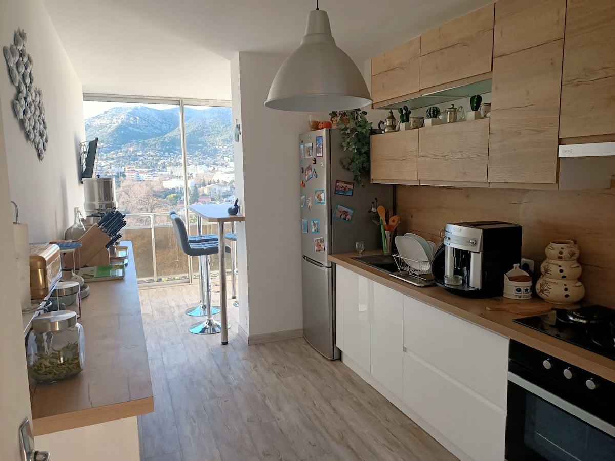 A modern kitchen is displayed with light wood cabinetry and white countertops. Appliances include a refrigerator, coffee maker, and stove. A tall glass table with stools is positioned next to a large window, offering views of the surrounding mountains and city below.