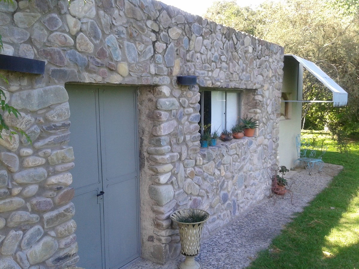 A stone exterior of the accommodation is displayed, characterized by a rustic design. Potted plants are seen in the window sill, and a covered porch extends over the entrance. A manicured lawn surrounds the structure, contributing to the natural setting.
