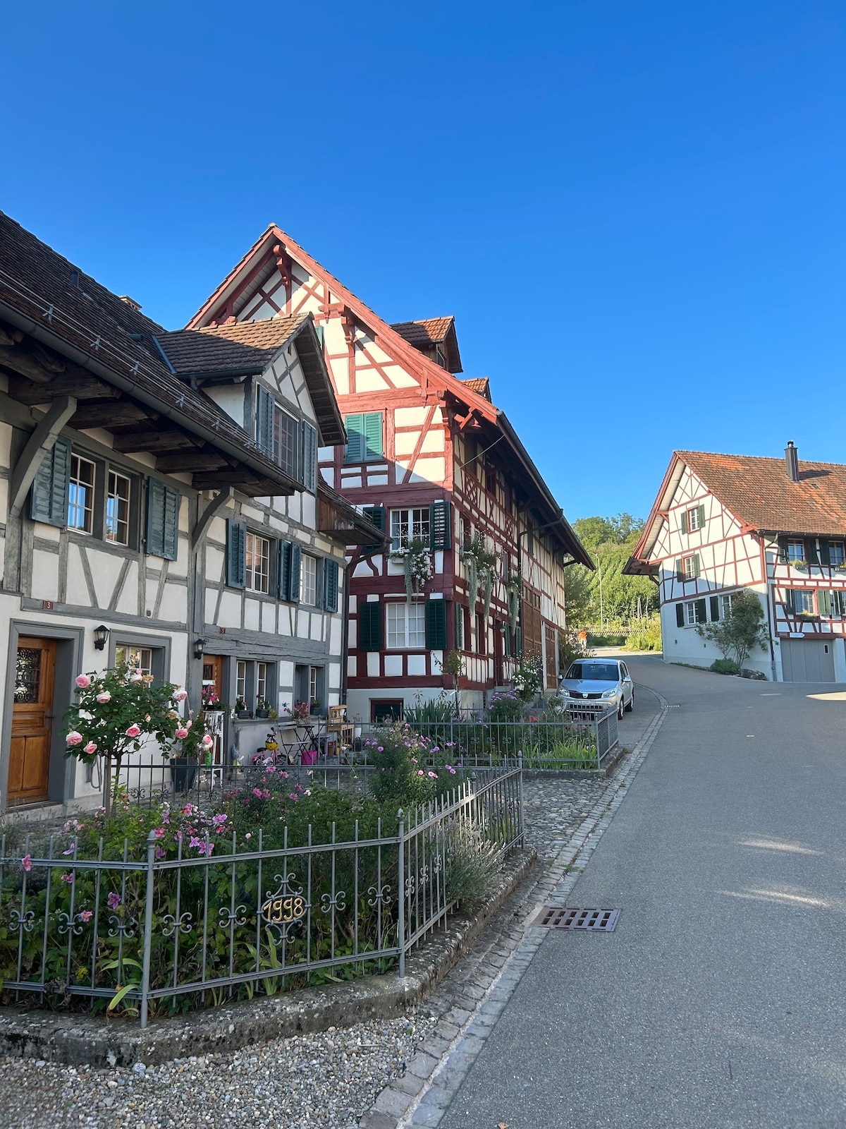 A charming streetscape features traditional Swiss half-timbered houses, showcasing unique architectural details. Flower beds and a decorative fence add to the inviting appearance. The surrounding area is quiet, with a clear blue sky visible overhead and a few vehicles parked along the road.