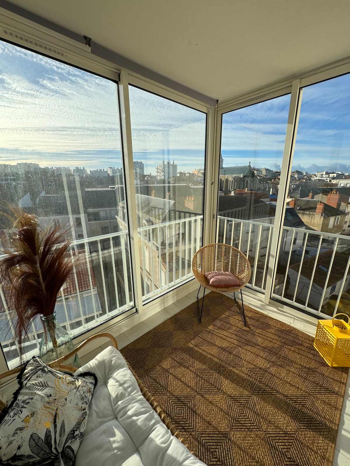 An open loggia features panoramic views of the city skyline, with sunlight streaming through large glass windows. A small, round chair sits beside a colorful cushion on a natural fiber rug, while decorative foliage adds a touch of greenery to the space.