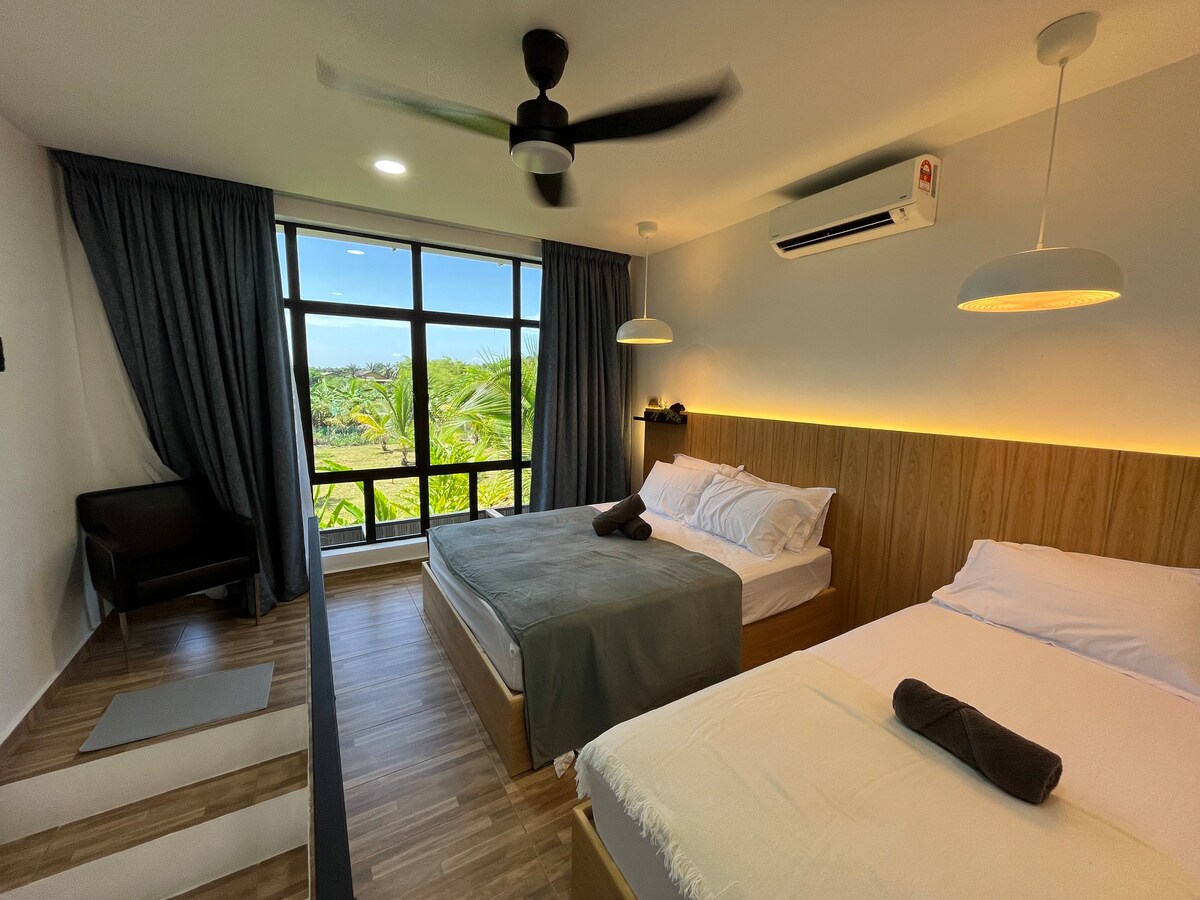 A bright and airy bedroom features two spacious beds, with soft linens and assorted pillows. Large windows allow natural light to fill the room, and curtains provide a contrast. A ceiling fan is installed for comfort, and a modest seating area is visible in the corner.