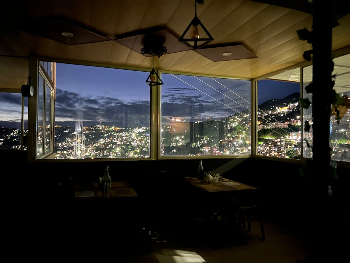 The dining area features large windows offering a panoramic view of illuminated Shimla at night. Soft lighting from ceiling fixtures highlights the wooden ceiling and table settings, creating a relaxed atmosphere. The distant hills are dotted with bright lights, providing a scenic backdrop.