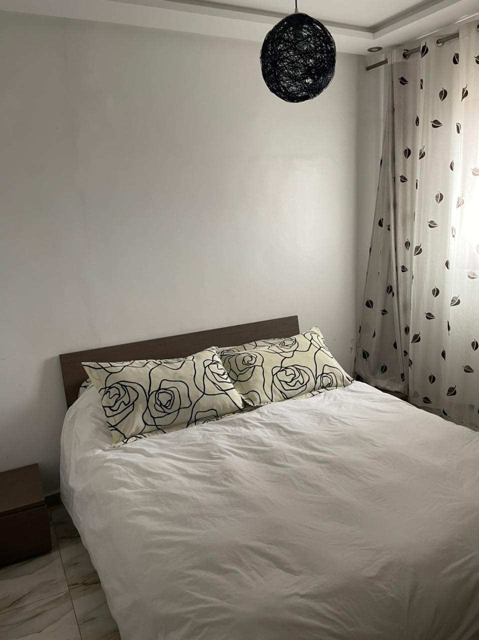 A light-filled bedroom features a spacious bed adorned with floral-patterned pillows. A simple bedside table is positioned beside the bed. Sheer curtains with a polka dot design allow natural light to filter into the room, while a round black light fixture hangs from the ceiling.