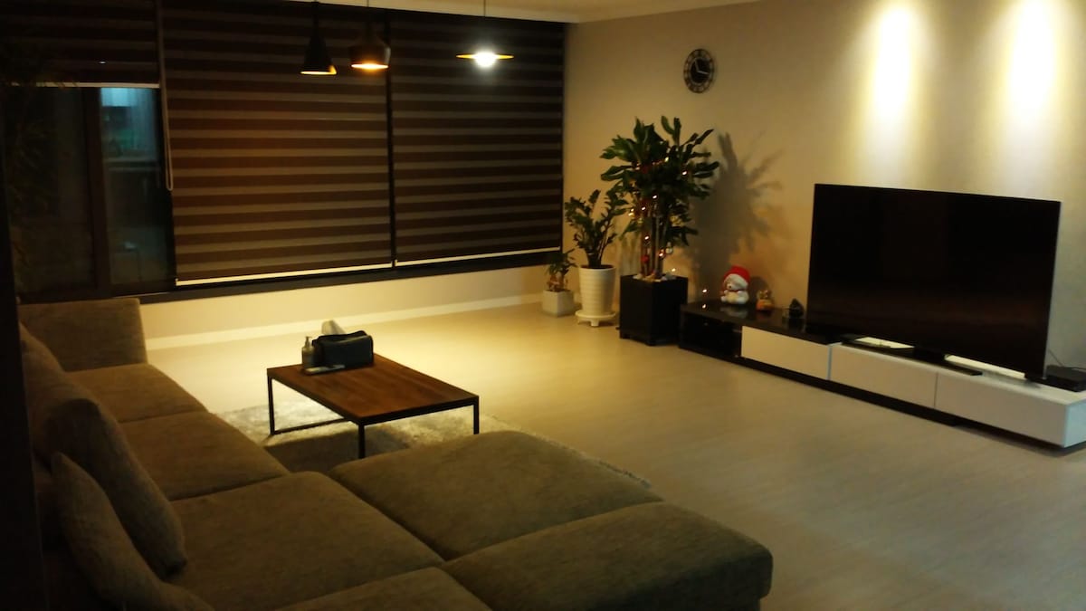 A spacious living area is adorned with a comfortable sectional sofa, a wooden coffee table, and a large flat-screen television. Tall plants add greenery to the space. Soft lighting is provided by overhead fixtures, and the room is complemented by layered dark window treatments.