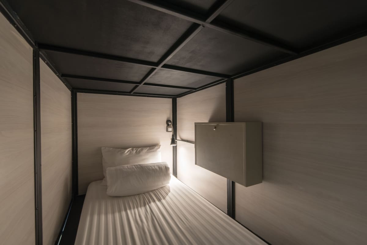 A cozy sleeping area is enclosed within wooden panels. A spacious bed is fitted with soft pillows and a striped duvet. A bedside lamp provides gentle illumination, while a small shelf is mounted on the wall for personal items.