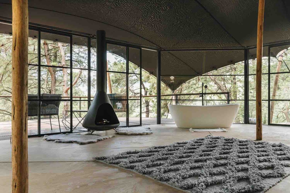 The interior space features a modern freestanding bathtub beside a black, sculptural fireplace. Large glass windows allow for ample natural light, offering views of the surrounding greenery. Textured rugs add warmth to the floor, complementing the unique architectural design.