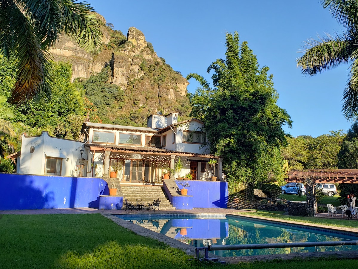 A spacious house with blue accents is nestled against a mountainous backdrop. The property features an inviting swimming pool surrounded by greenery, with outdoor seating provided. The facade showcases a combination of rustic design and modern amenities, reflecting a harmonious connection with nature.