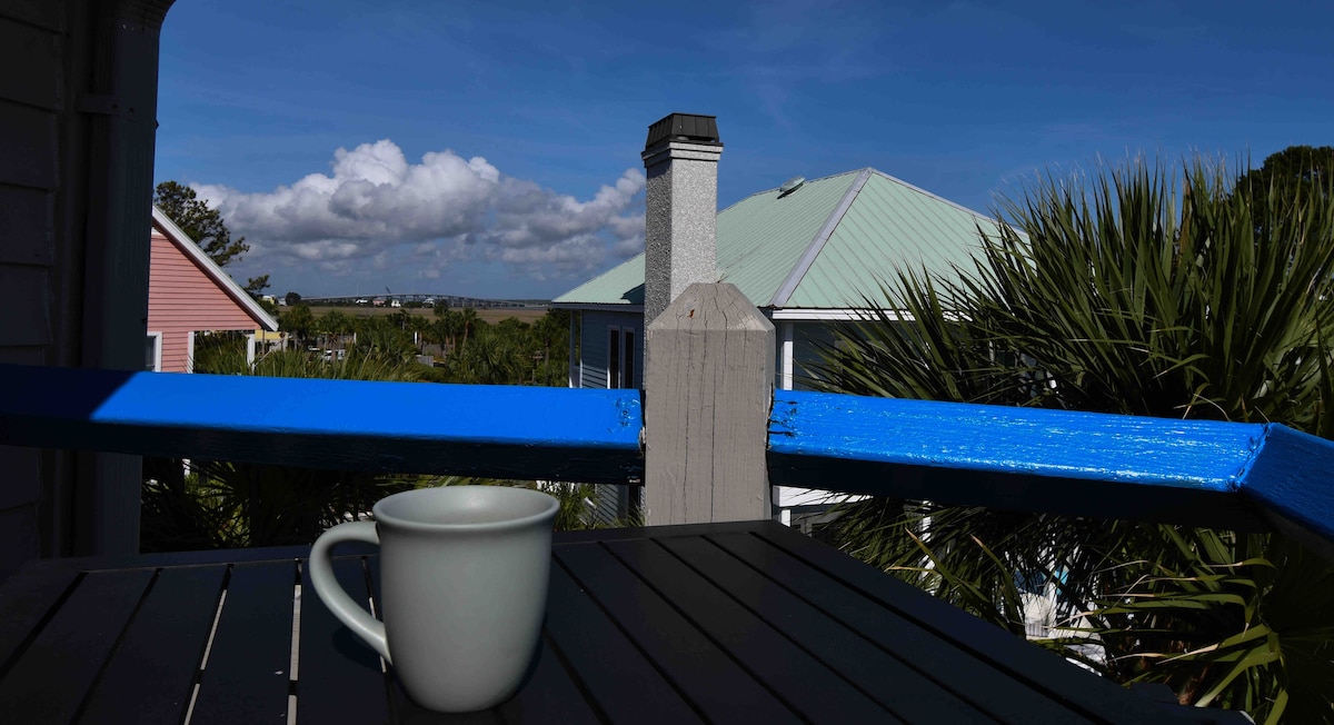 A light blue balcony railing frames a table, where a pale green cup rests. In the background, the view includes rooftops with green and pink accents, surrounded by lush palm trees under a clear sky with scattered clouds.