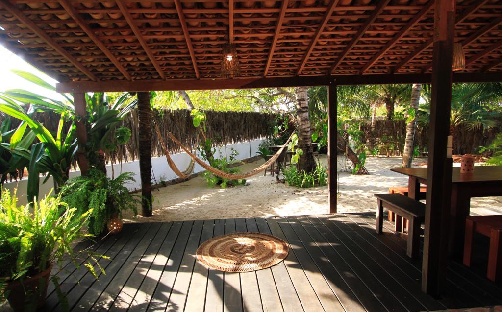 An open terrace is shown with wooden flooring and a thatched roof. A hammock is visible between two palm trees, offering a serene place to relax. Lush greenery surrounds the area, enhancing the tropical feel of the garden. Natural light filters through the overhead structure.
