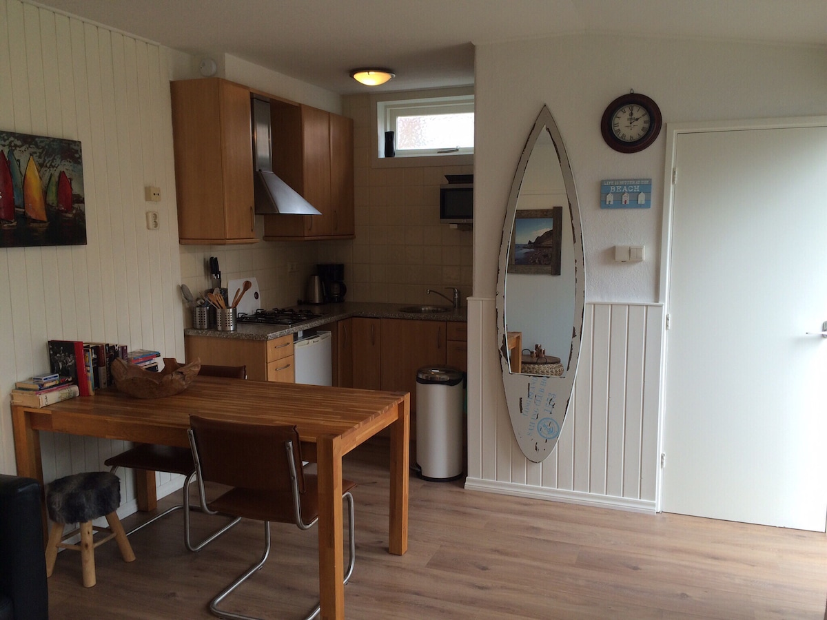 A functional kitchen area features wooden cabinets and a stainless-steel stove. A small dining table is set with two chairs, and a mirror shaped like a surfboard is mounted on the wall. A clock hangs above the kitchen space, contributing to a clean and organized look.