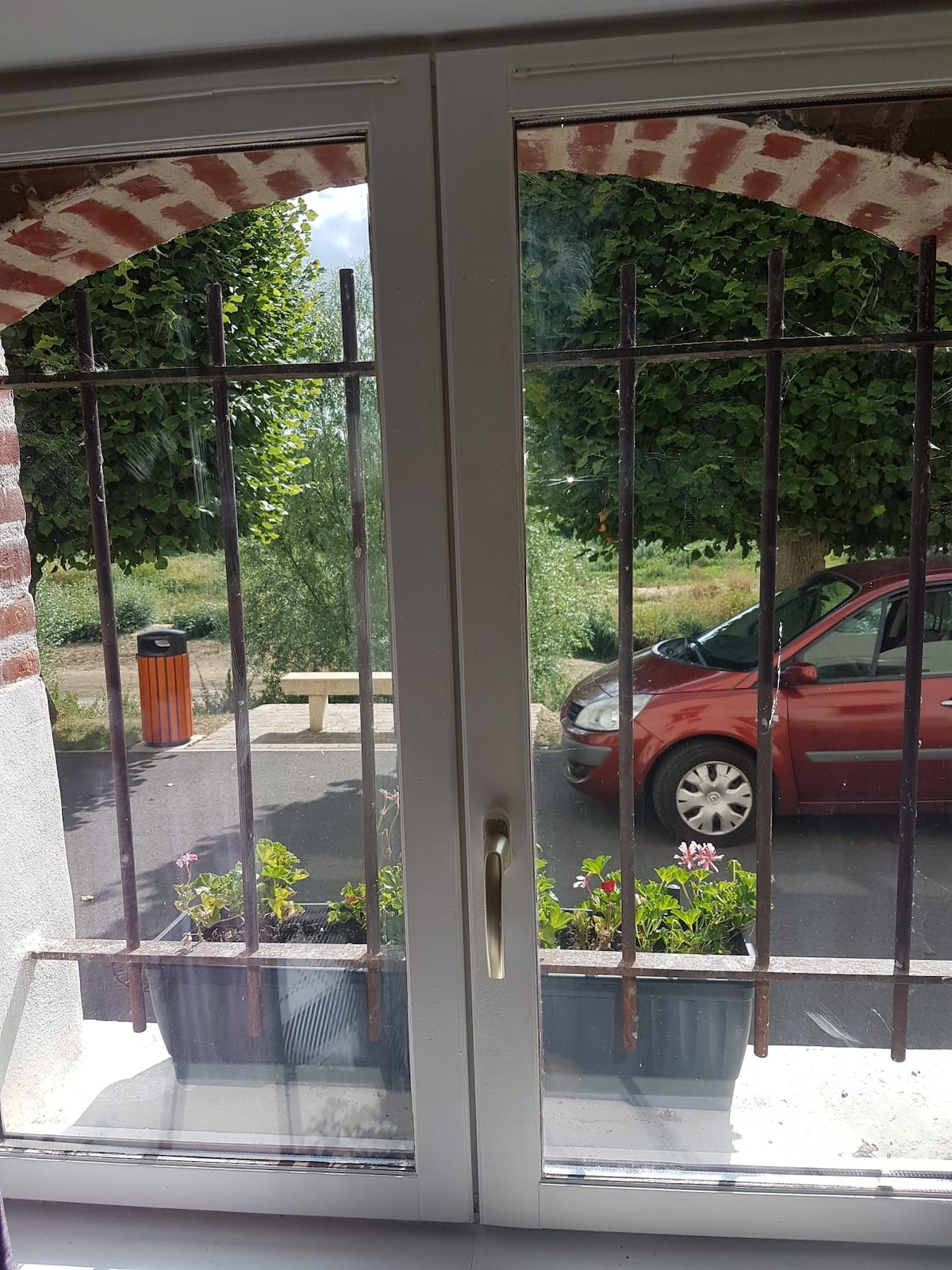 A view from a window features bars for security, framing a glimpse of the outside street. Potted flowers rest on the windowsill, while a red car is parked nearby. Green trees line the street, contributing to a serene environment.