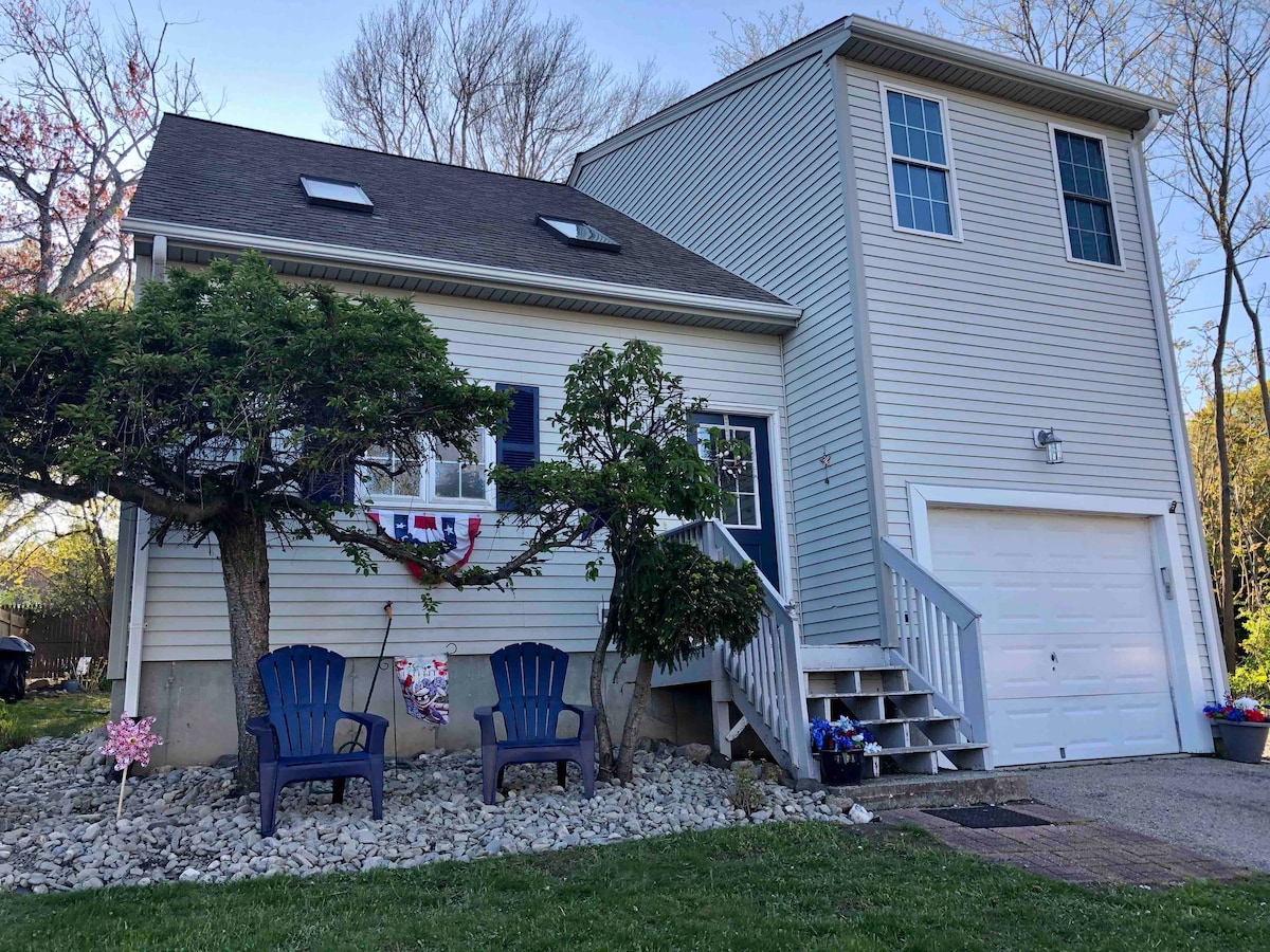 The exterior of a coastal home is shown, featuring light-colored siding and a sloped roof. Two blue Adirondack chairs are positioned on a stony area in front, with a staircase leading to the entrance. Flower pots add a touch of color to the landscape.