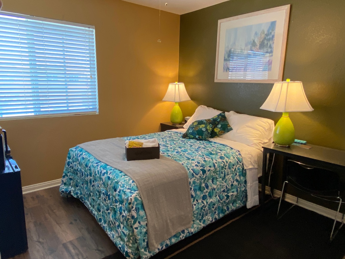 A cozy bedroom is furnished with a spacious bed covered in vibrant blue and green bedding. Two lamps with green bases provide soft lighting, while a piece of artwork hangs above the bed. A window featuring blinds allows natural light to filter in.