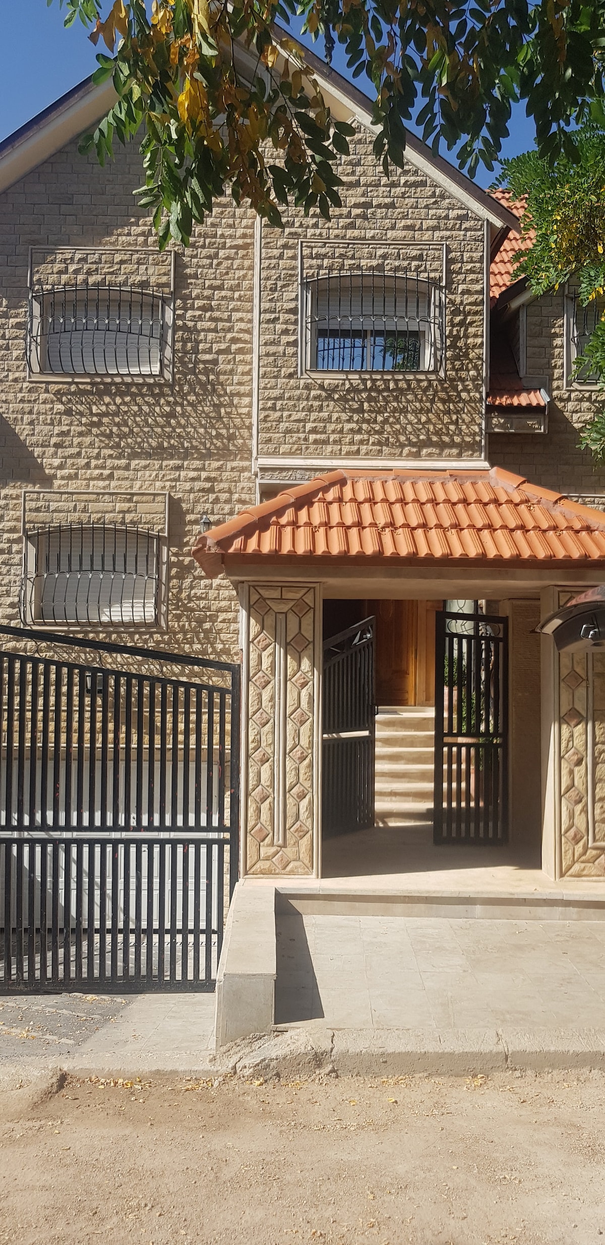The exterior of a spacious villa features a stone facade and a red-tiled roof. A gated entrance leads to stairs ascending to the main entrance, surrounded by greenery. Windows with decorative bars allow natural light into the interior.