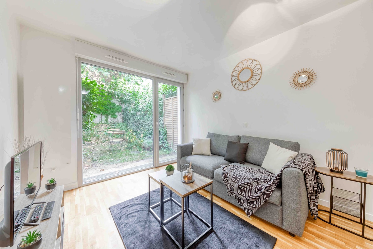 A bright living area features a spacious grey sofa, a coffee table, and a television placed on a low console. Large glass doors open to a private garden, enhancing natural light. Decorative mirrors adorn the walls, complemented by a plush rug underfoot.