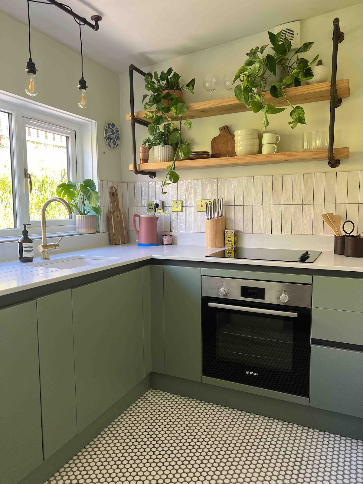 The kitchen features modern cabinetry in soft green tones, complemented by open shelving adorned with potted plants and dishware. A sleek oven and a contemporary sink are positioned under a window, allowing natural light to brighten the space, which showcases a patterned tile floor.