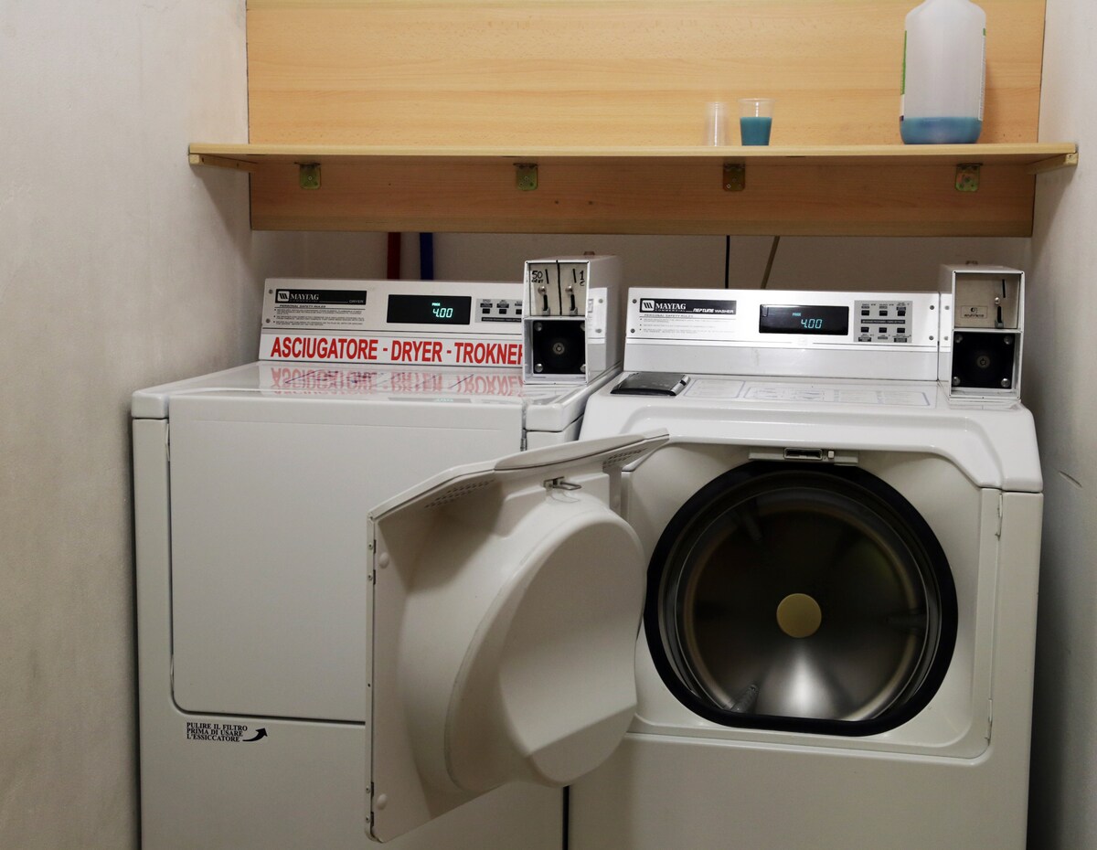 A self-service laundry area is shown, featuring a washing machine and a dryer. The machines are white and equipped with digital controls. A shelf above holds a container and a cup. Clear labels indicate their functions, providing convenient laundry options for guests.