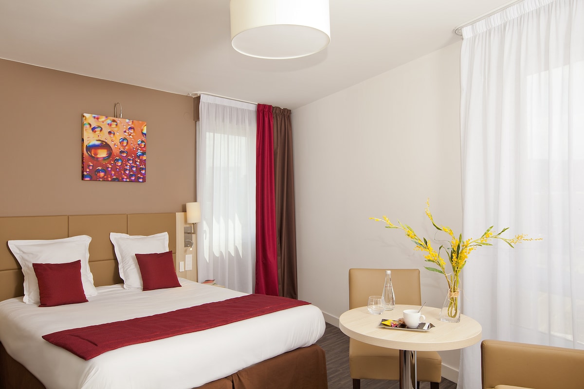 A well-lit studio features a spacious bed with white linens and burgundy accents. A small round table is set with a vase of yellow flowers, accompanied by two beige chairs. Bright curtains frame large windows, allowing natural light to fill the space.