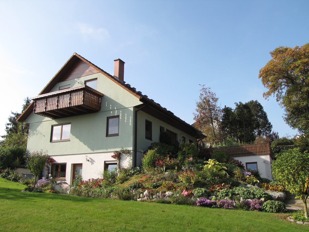A two-story house is set in a lush garden, featuring a balcony on the upper level. The exterior is surrounded by vibrant flowers and greenery, with a clear blue sky overhead. The structure's design highlights a blend of natural beauty and inviting architecture.