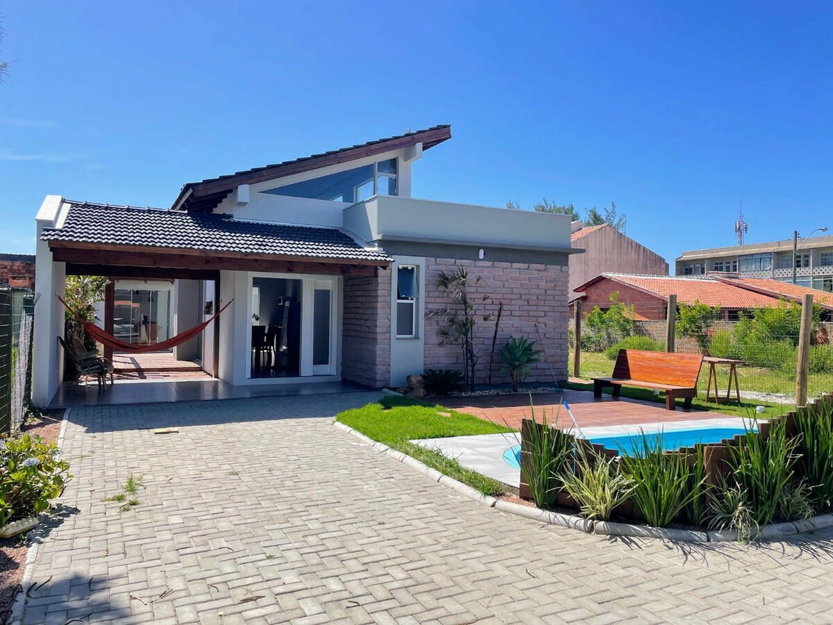A modern house showcases a well-maintained exterior with a paved pathway and a manicured lawn. Adjacent to the house, a small pool and a wooden deck are visible, inviting relaxation. A hammock is hung between posts, offering a cozy spot for leisure.