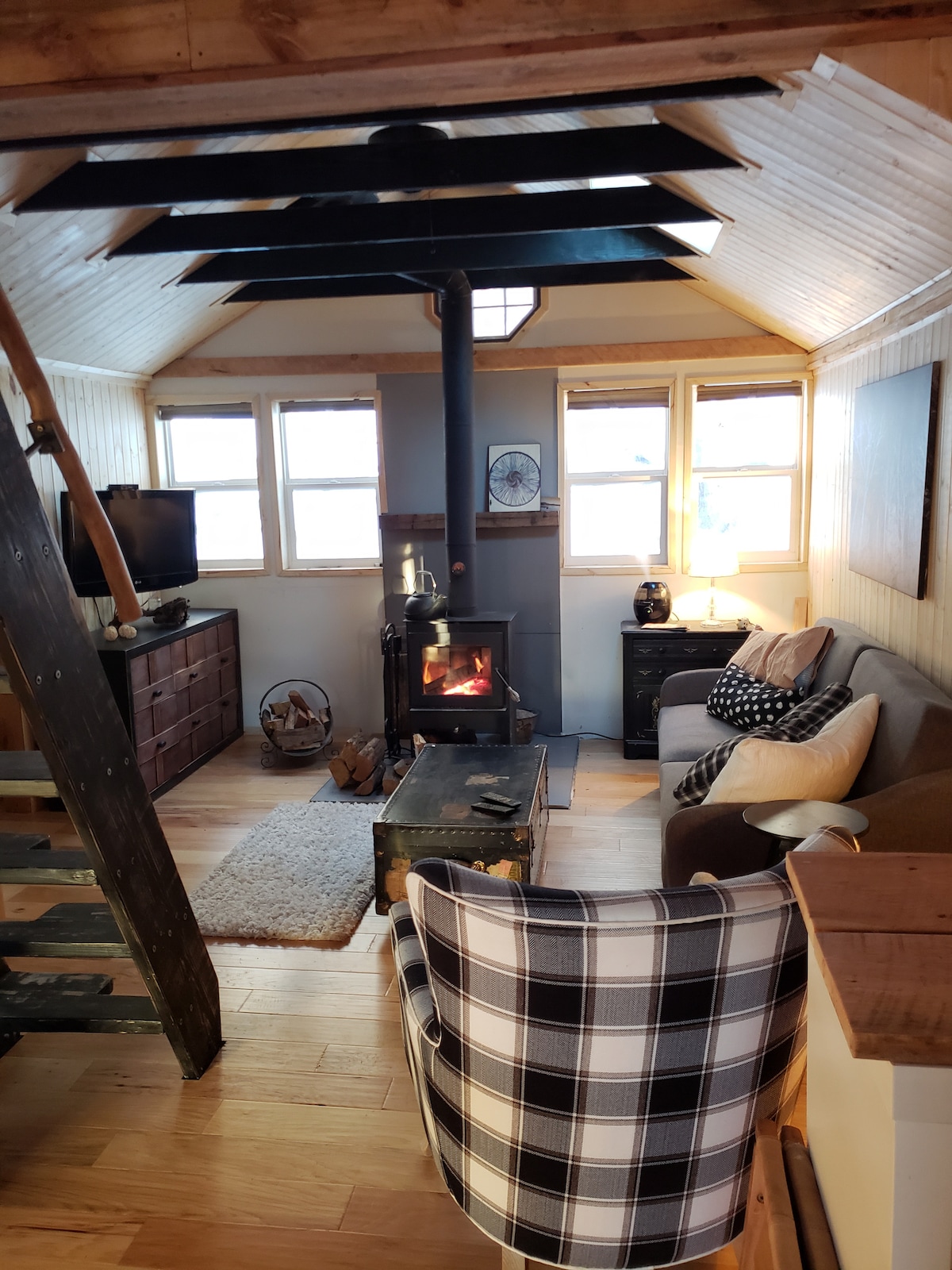A cozy living space features a comfortable sofa and an armchair with a checkered pattern. Natural light streams through multiple windows, highlighting a small wood stove at the center. A wooden staircase leads to the loft, and a TV is positioned on a low dresser nearby.