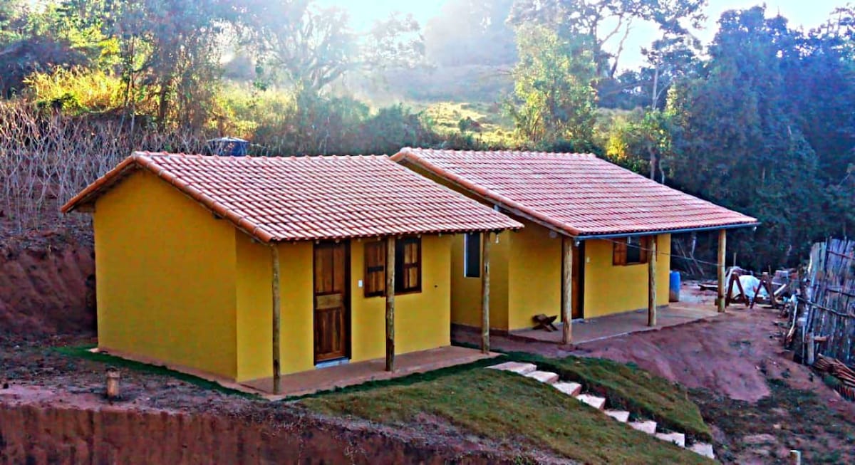 Two charming yellow chalets are set on a hillside, surrounded by green hills. Each building features a tiled roof and wooden doors. A stone pathway leads up to the entrance, while the natural landscape provides a serene backdrop.