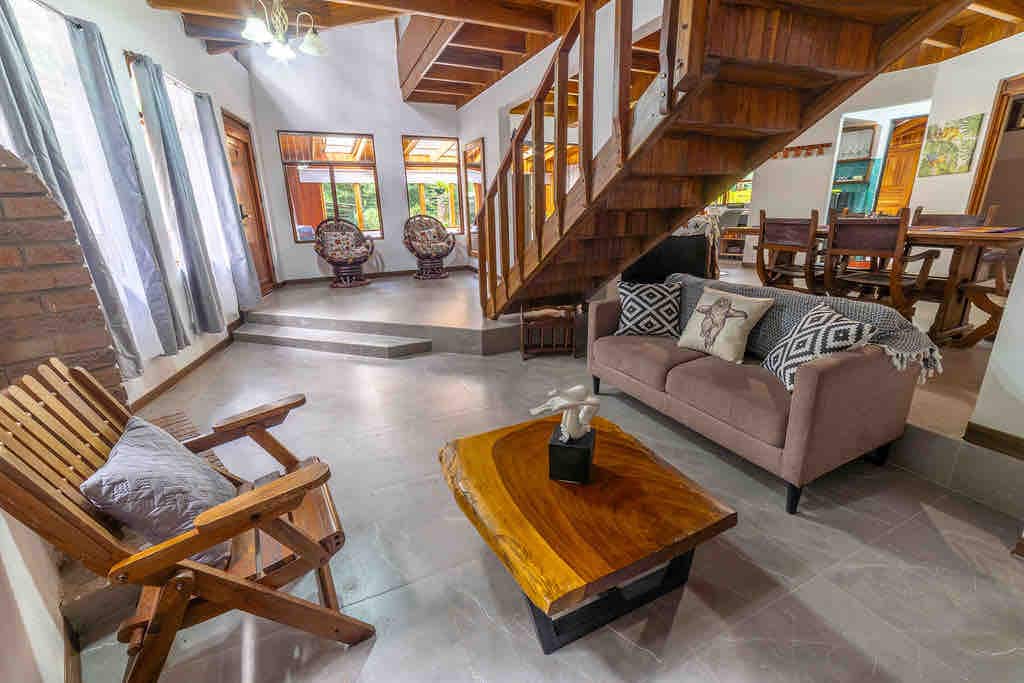 A spacious living area is shown, featuring natural wood accents and an open floor plan. A comfortable sofa is complemented by a unique wooden coffee table, while two chairs add additional seating. Large windows allow ample natural light, casting a warm glow over the space.