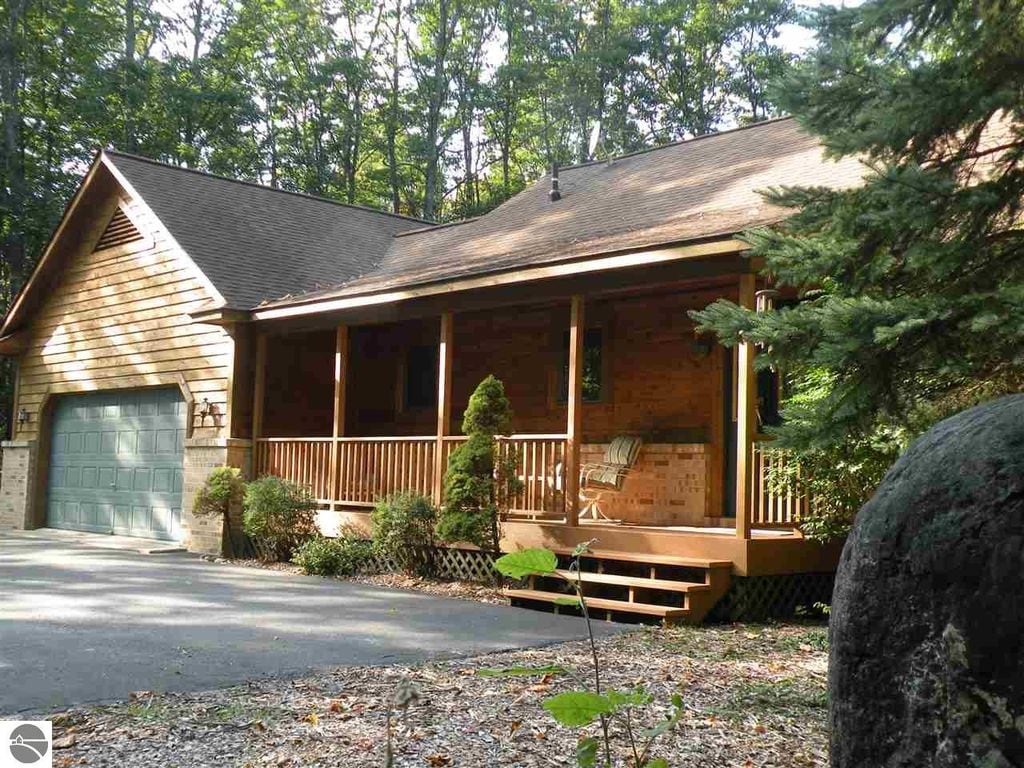 A spacious wooden cabin is nestled among tall trees, featuring a covered porch with two rocking chairs. The garage door is visible on the left, and a stone pathway leads to the entrance. Natural surroundings enhance the cabin's rustic charm.