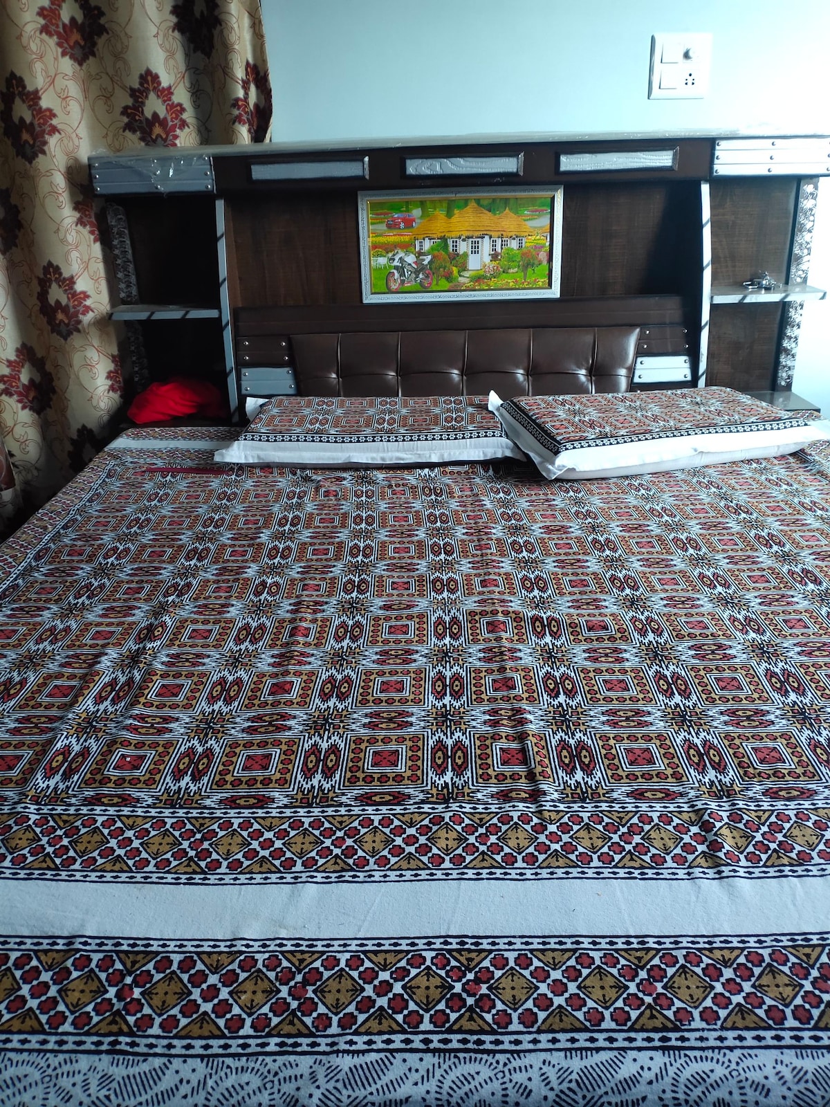 A spacious bed is centered in the room, adorned with a patterned bedspread in earthy tones. A decorative framed artwork hangs above the headboard. Wooden shelves flank the bed, holding items within reach. Soft curtains frame a nearby window, adding a touch of warmth.