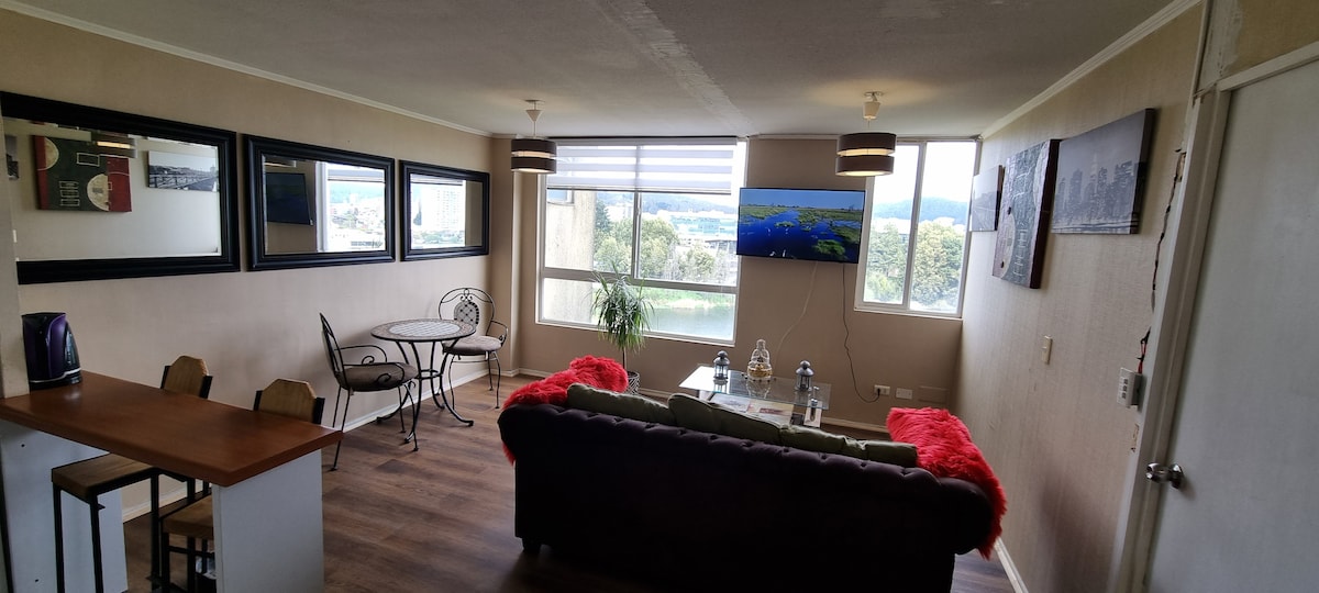 A cozy living space features a dark sofa with red accents, paired with a glass coffee table. Large windows offer a view of the surroundings. A dining area with two chairs and a small round table is situated nearby, along with a well-equipped kitchen area.