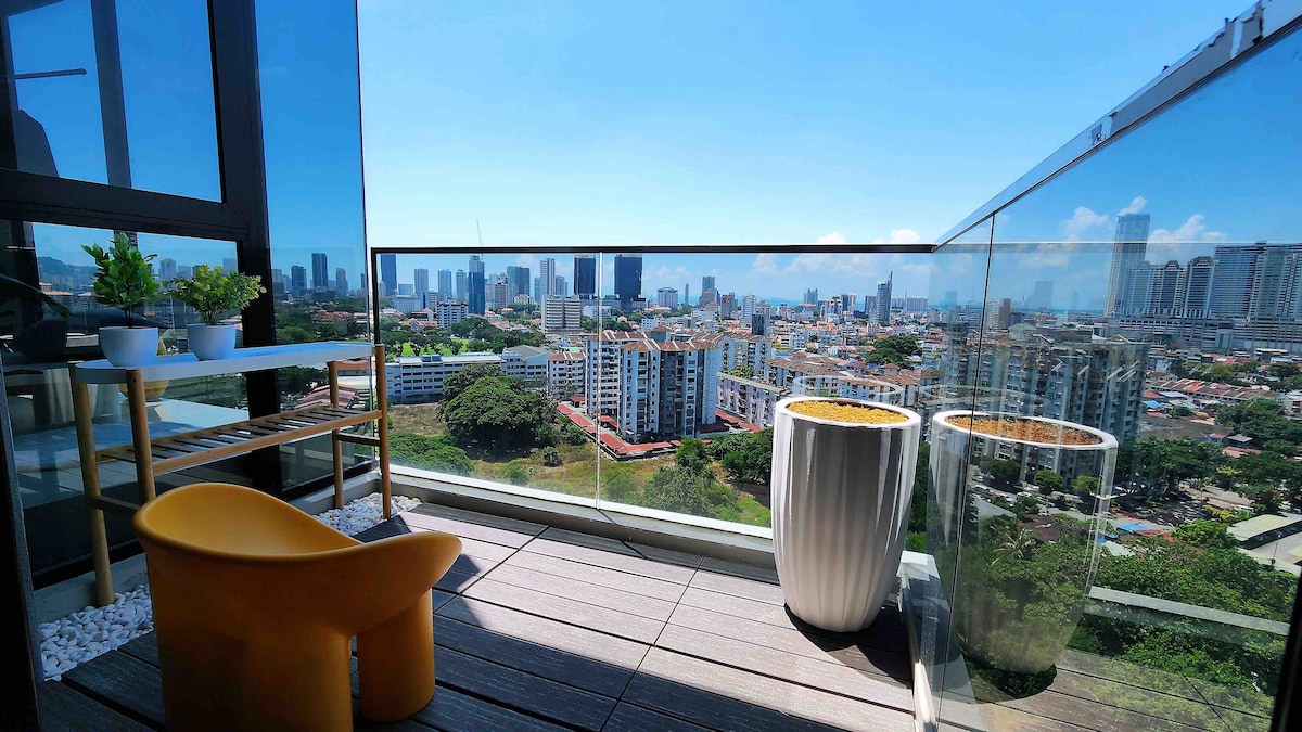 A spacious balcony provides panoramic views of the city skyline. Two modern planters and a small wooden table are visible, complemented by a bright yellow chair. Sunlight illuminates the area, enhancing the sense of outdoor relaxation.