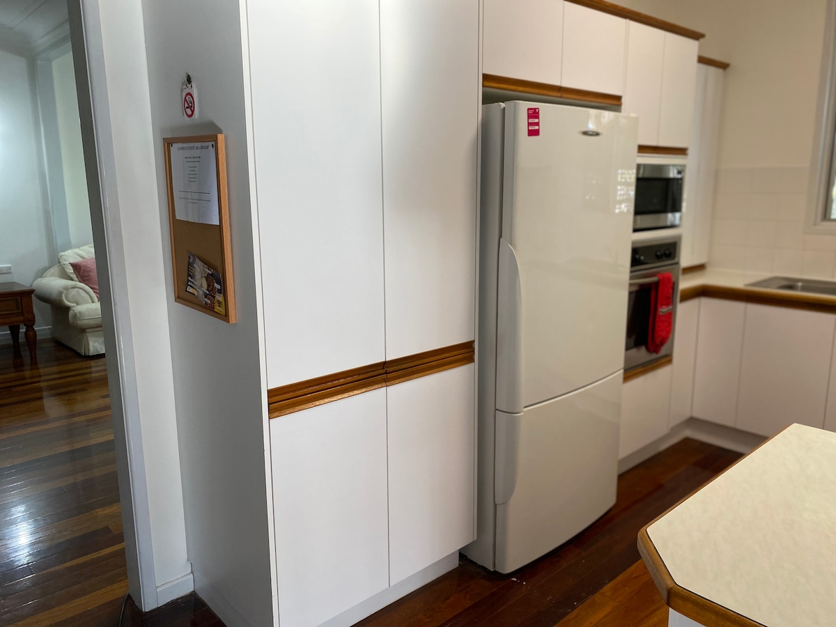 A modern kitchen featuring white cabinetry and wooden accents. A stainless steel refrigerator is positioned next to an oven and microwave unit built into the cabinetry. The kitchen floor is adorned with polished wooden planks, contributing to the overall clean and functional design.