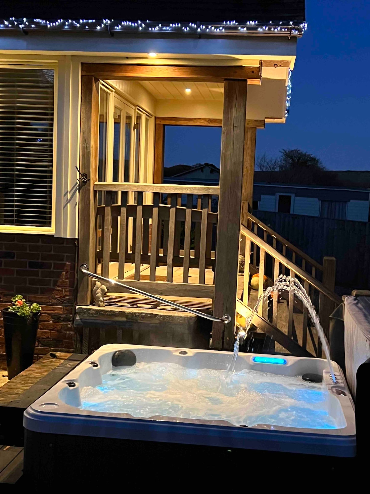 A hot tub is positioned on a wooden deck, with water gently flowing from the spouts. Soft blue lighting emanates from the tub, while the exterior staircase leads towards a bright entrance. Twinkling lights adorn the overhead area, enhancing the serene evening setting.