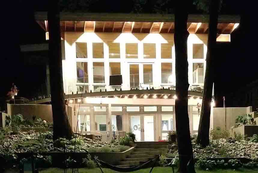 A modern home is illuminated at night, showcasing large windows that reveal the interior. The structure is flanked by trees, and stairs lead to a landscaped entrance surrounded by flowers. Soft lighting enhances the architectural details and provides a welcoming ambiance.
