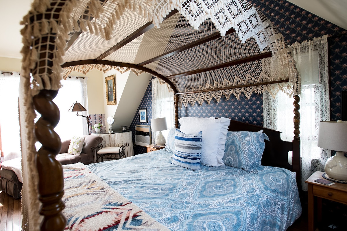 A canopied bed features a blue and white patterned comforter, complemented by decorative pillows. Soft light enters through sheer curtains, illuminating the cozy space. A sitting area with a chair and lamp is visible in the corner, enhancing the room's inviting atmosphere.