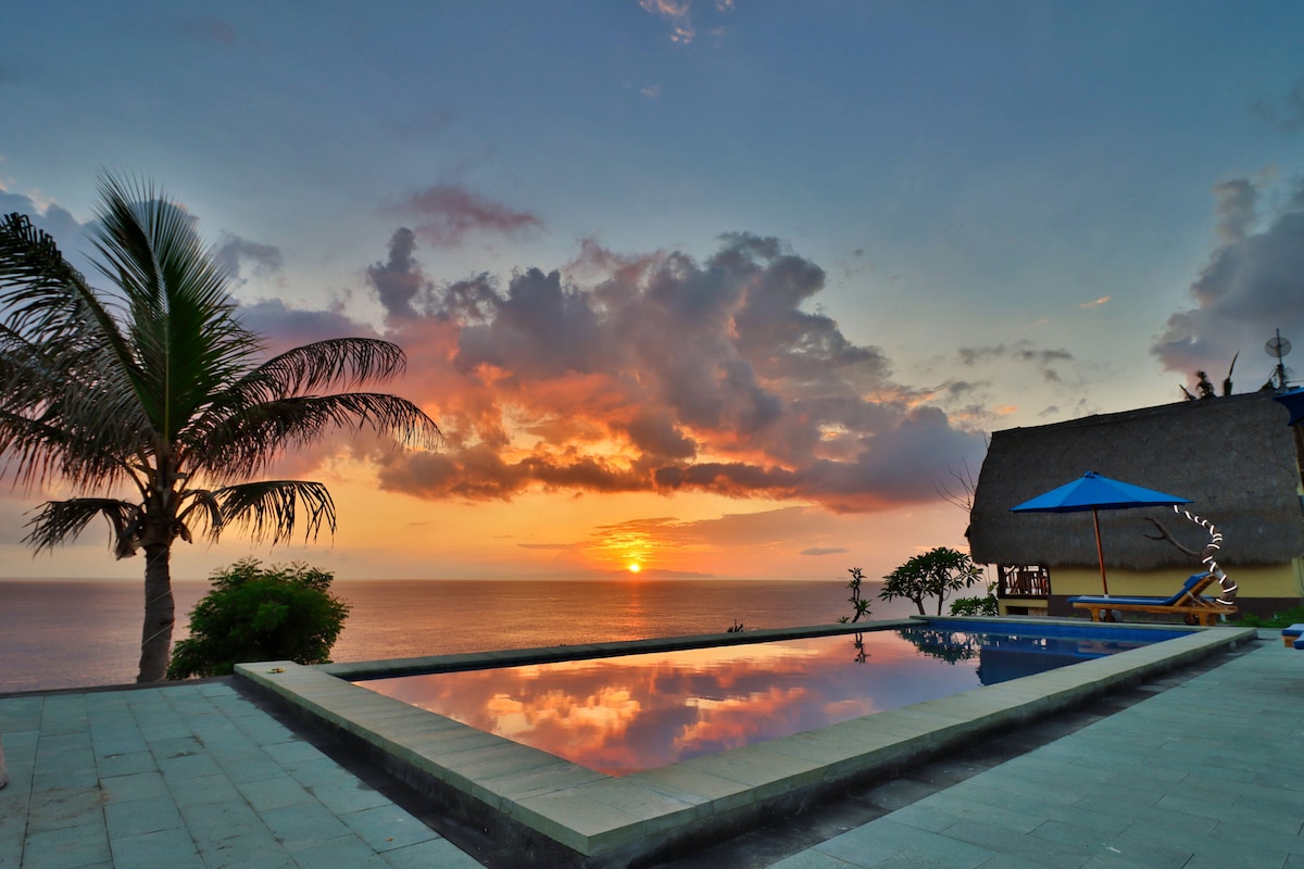 A tranquil infinity pool reflects a vibrant sunset over the ocean, framed by palm trees. The colors of the sky blend softly as the sun descends on the horizon, highlighting the serene ambiance of the outdoor space.