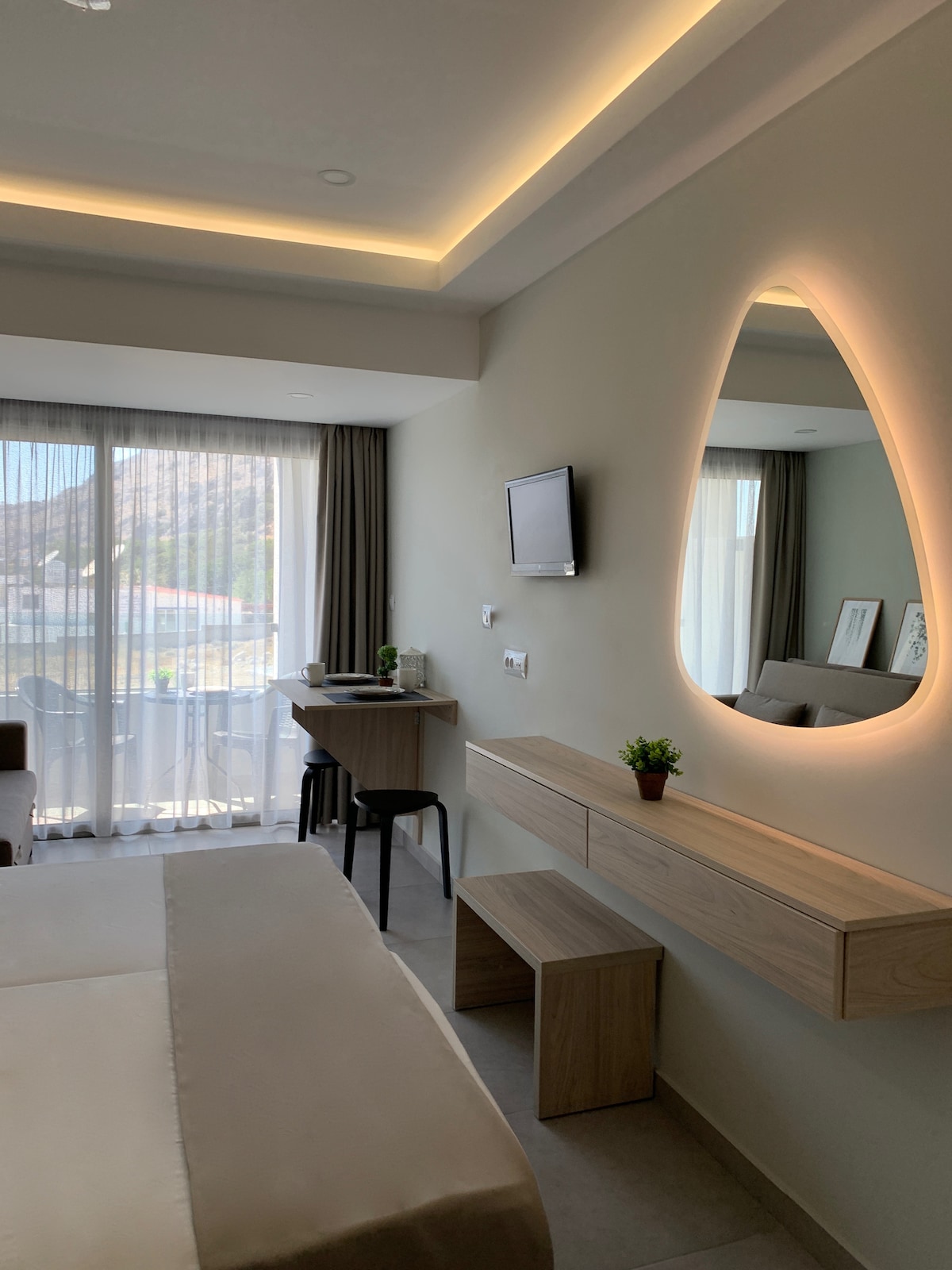 A modern studio features a spacious bed with soft linens against a neutral-toned wall. A stylish mirror hangs above a wooden console, complementing the light wood furniture and a small workspace. Natural light fills the room through glass doors leading to the private balcony.