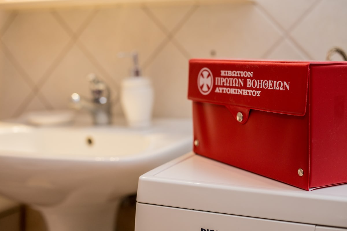 A red first aid kit is placed on top of a white appliance in a bathroom setting, with a sink visible in the background. Soft lighting creates a clean and organized atmosphere, emphasizing the kit's accessibility for any emergencies.